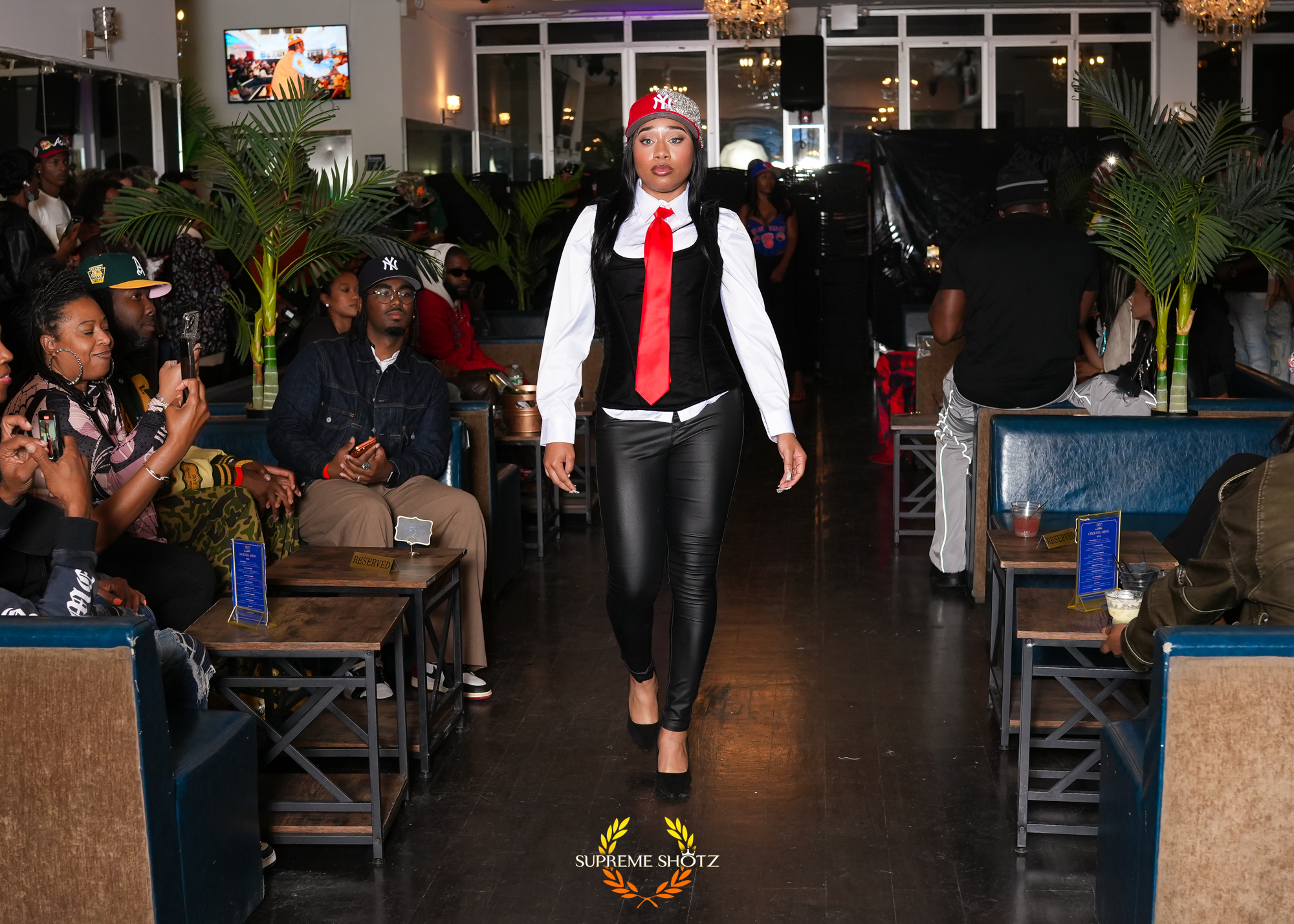 Taniya walking the runway in a black corset vest, white shirt, red tie and leather pants
