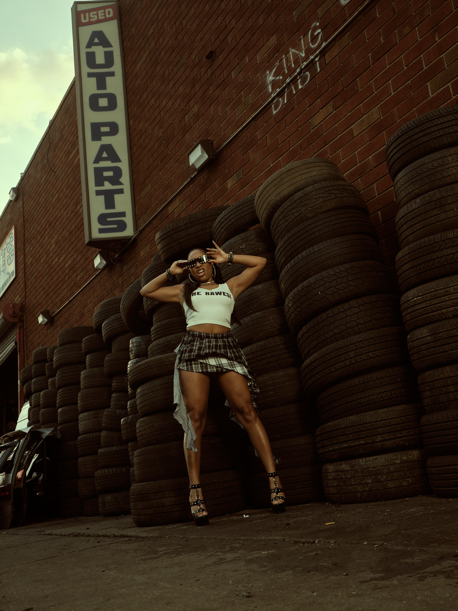 Low-angle shot of Taniya under an Auto Parts sign surrounded by stacked tires