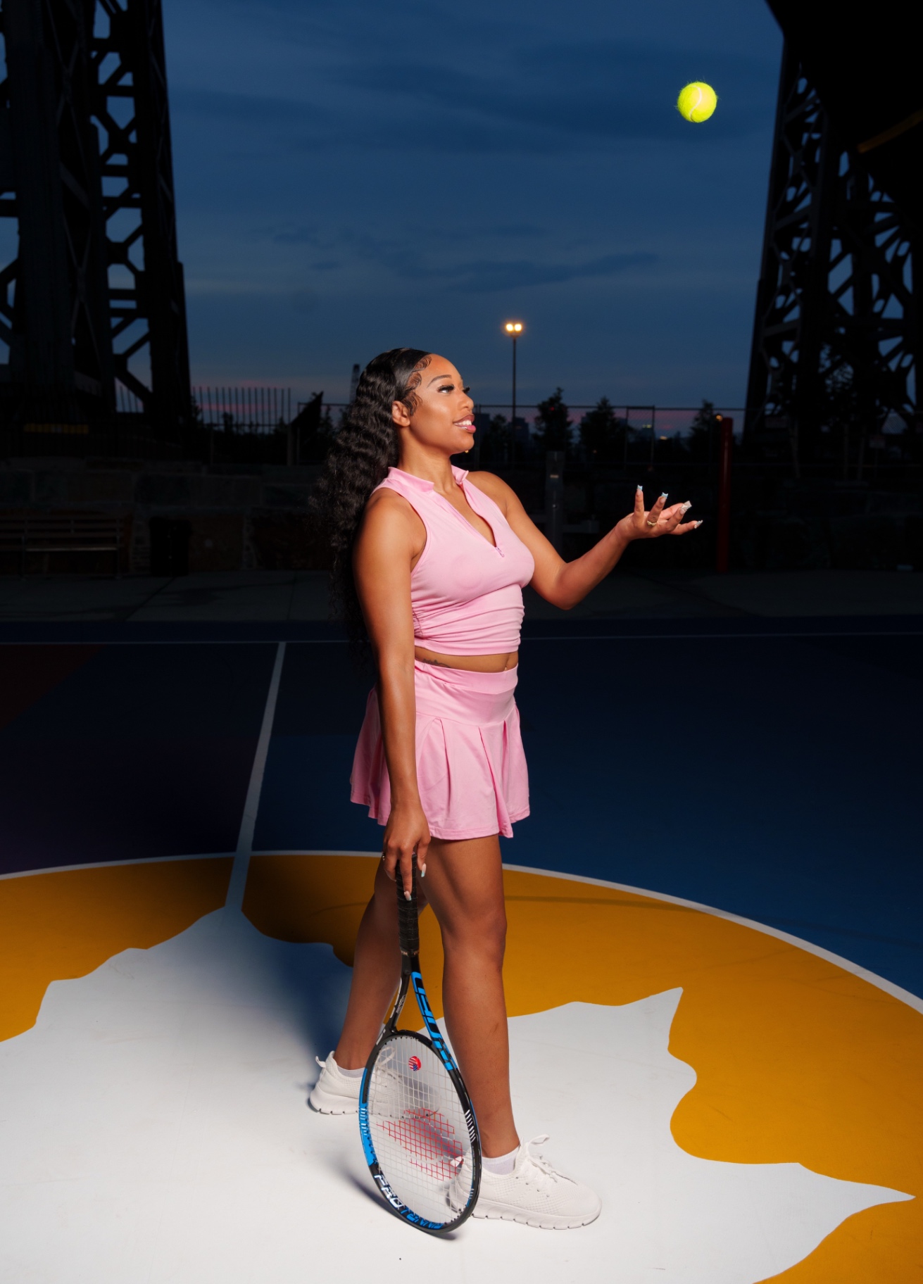Taniya tossing a tennis ball on a court at dusk in a pink tennis set, city bridge behind her