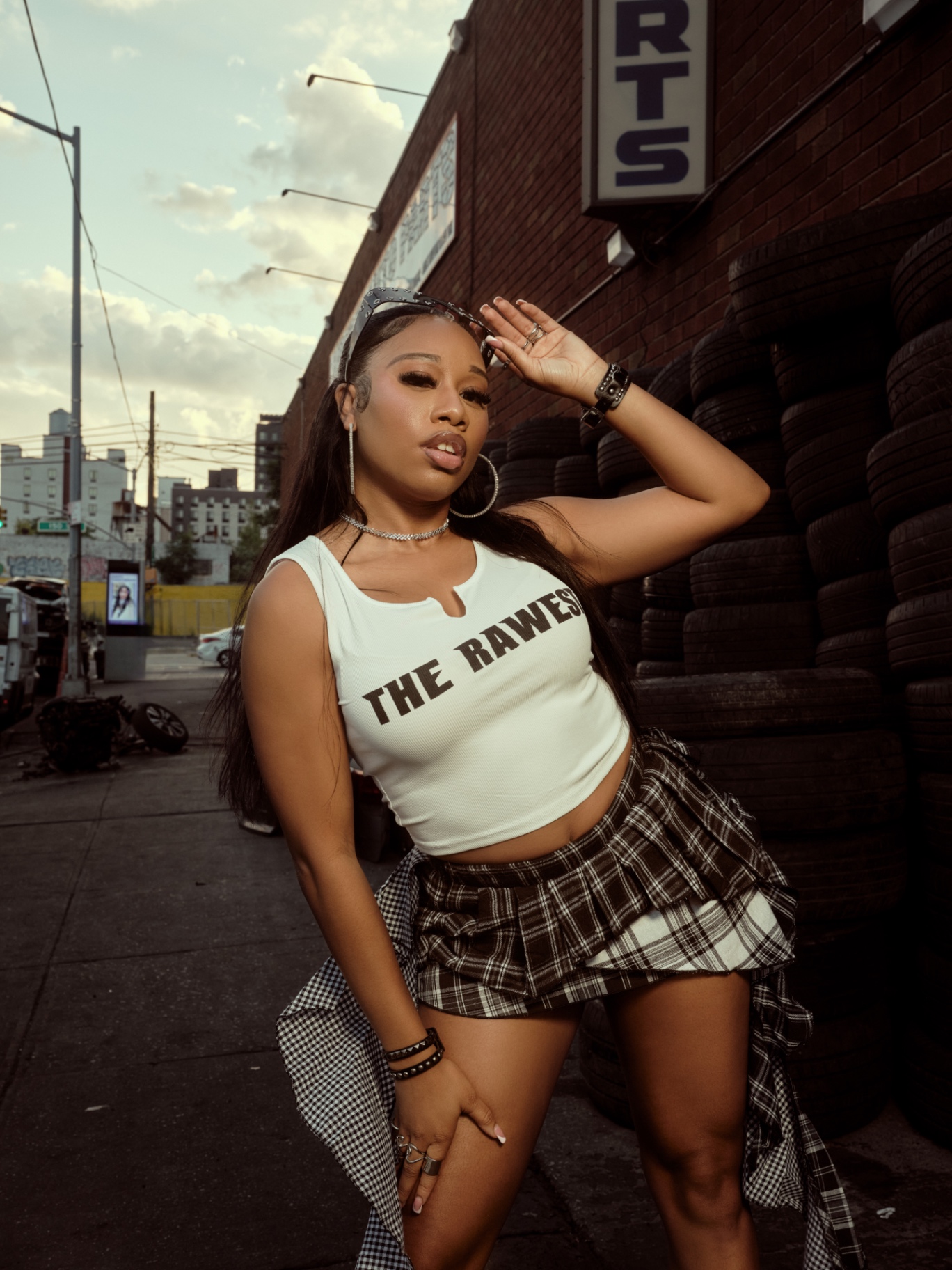 Taniya posing against a brick auto-parts wall with stacked tires at golden hour