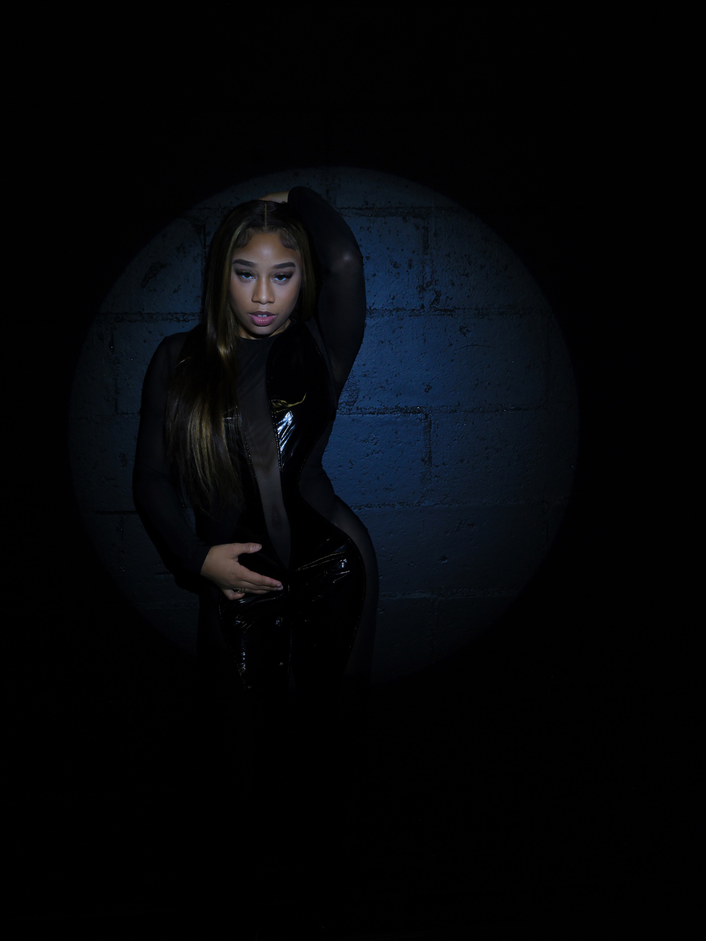 Taniya in a black mesh and patent bodysuit lit by a single spotlight against a blue wall