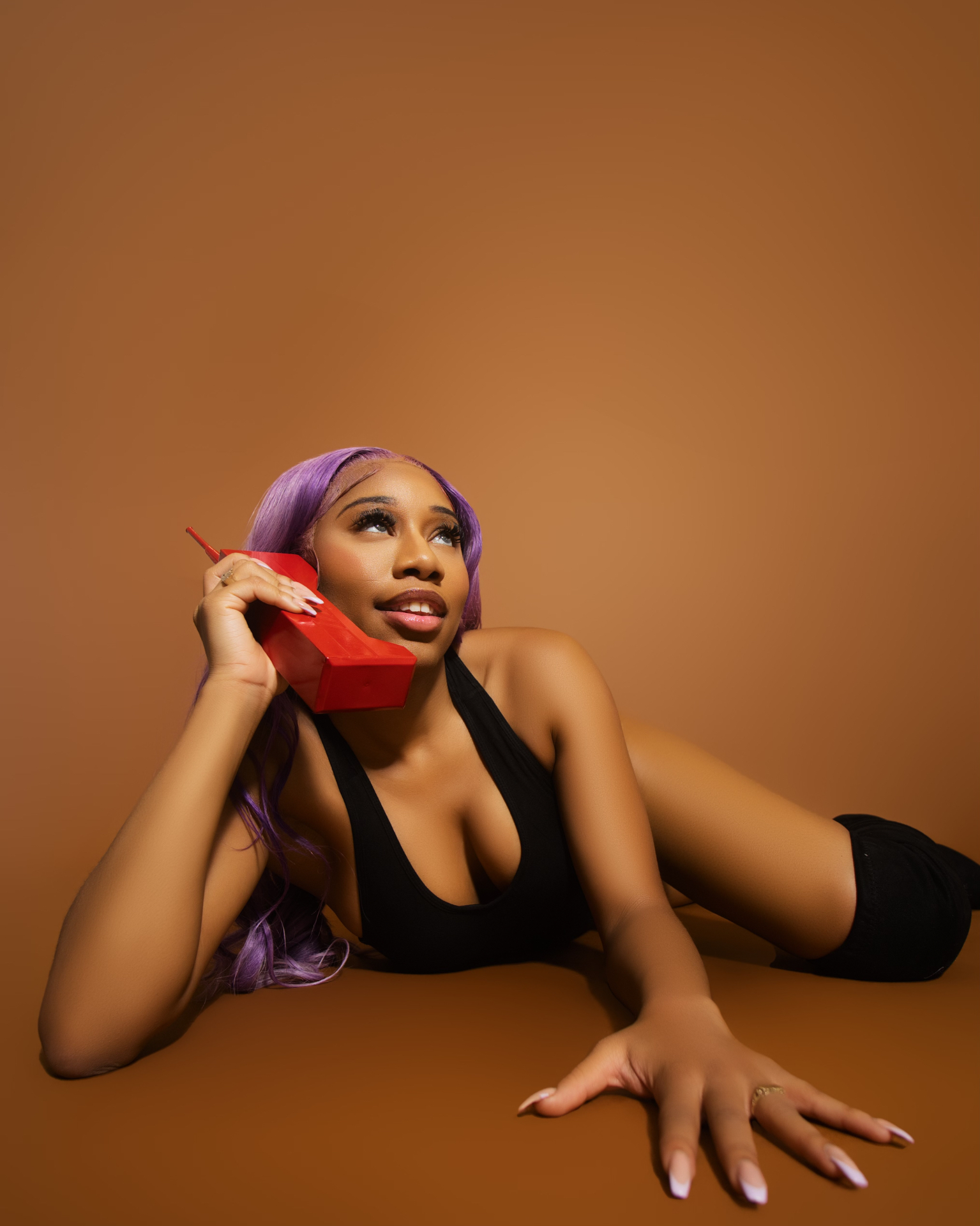 Taniya with lavender hair reclining on a caramel backdrop with a retro red phone