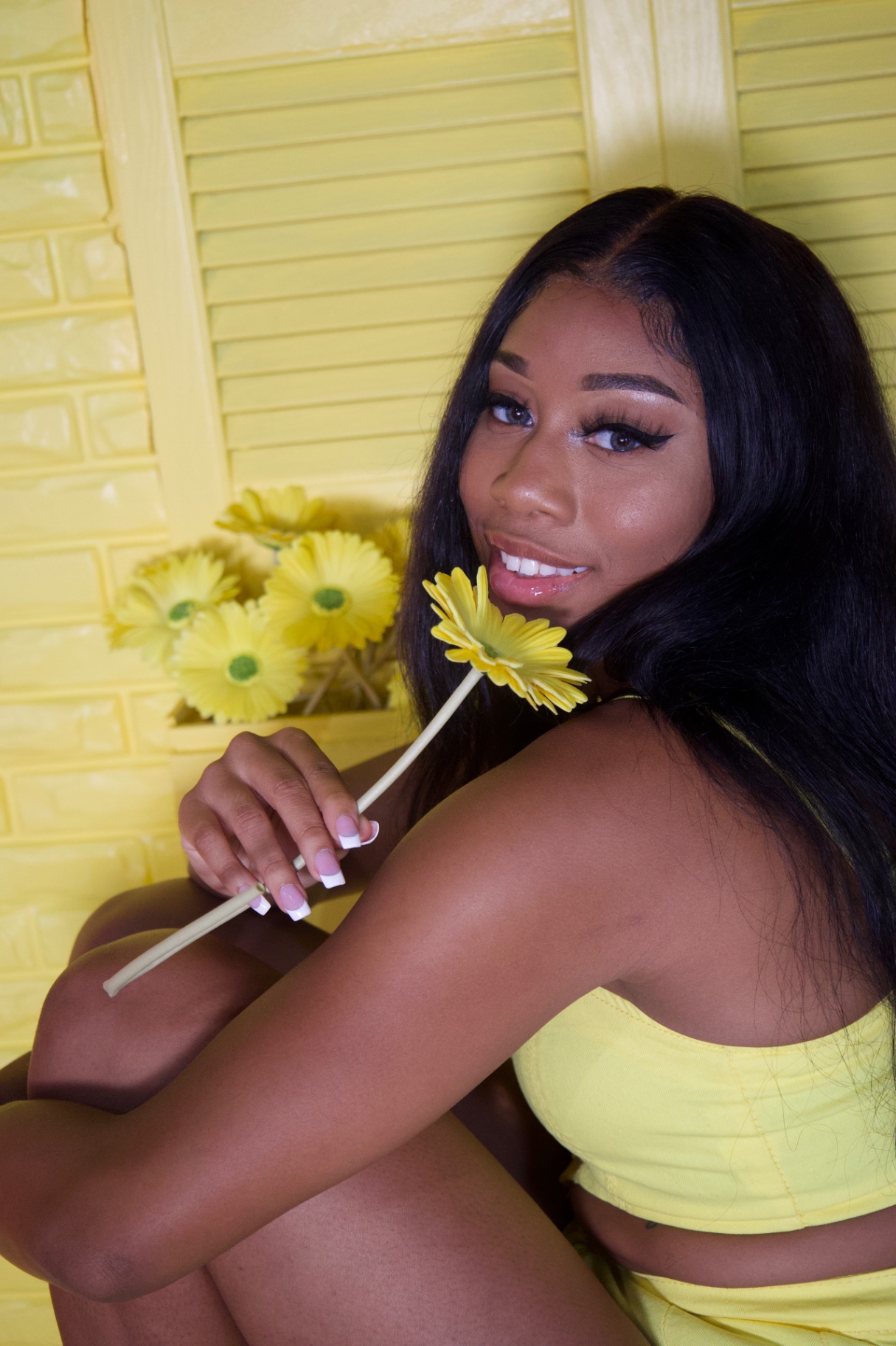 Taniya seated in a yellow two-piece holding a yellow gerbera daisy against a yellow set