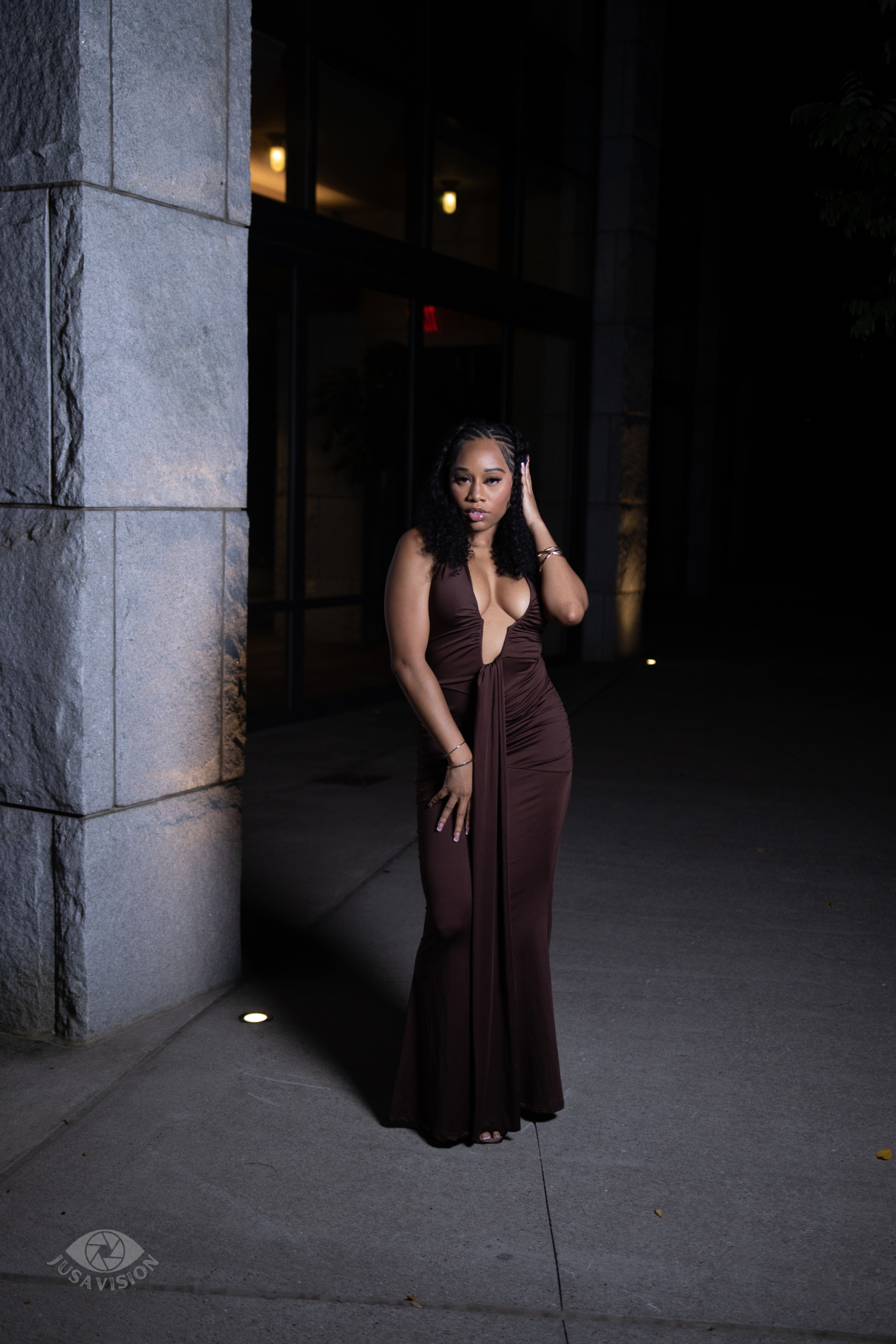 Taniya in a chocolate brown gown with one hand in her hair, night city lighting