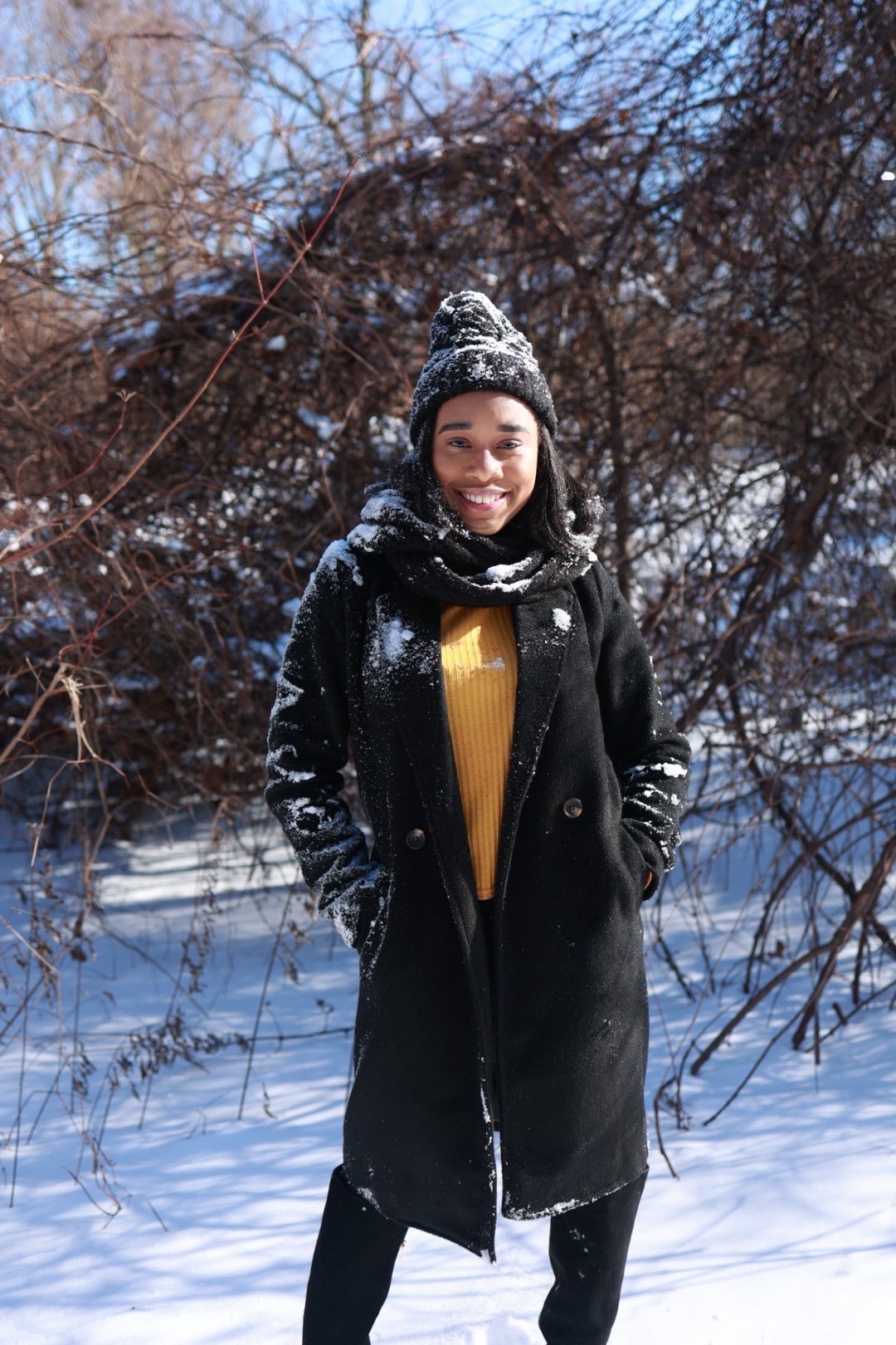 Taniya smiling in a long black coat and beanie standing in deep snow