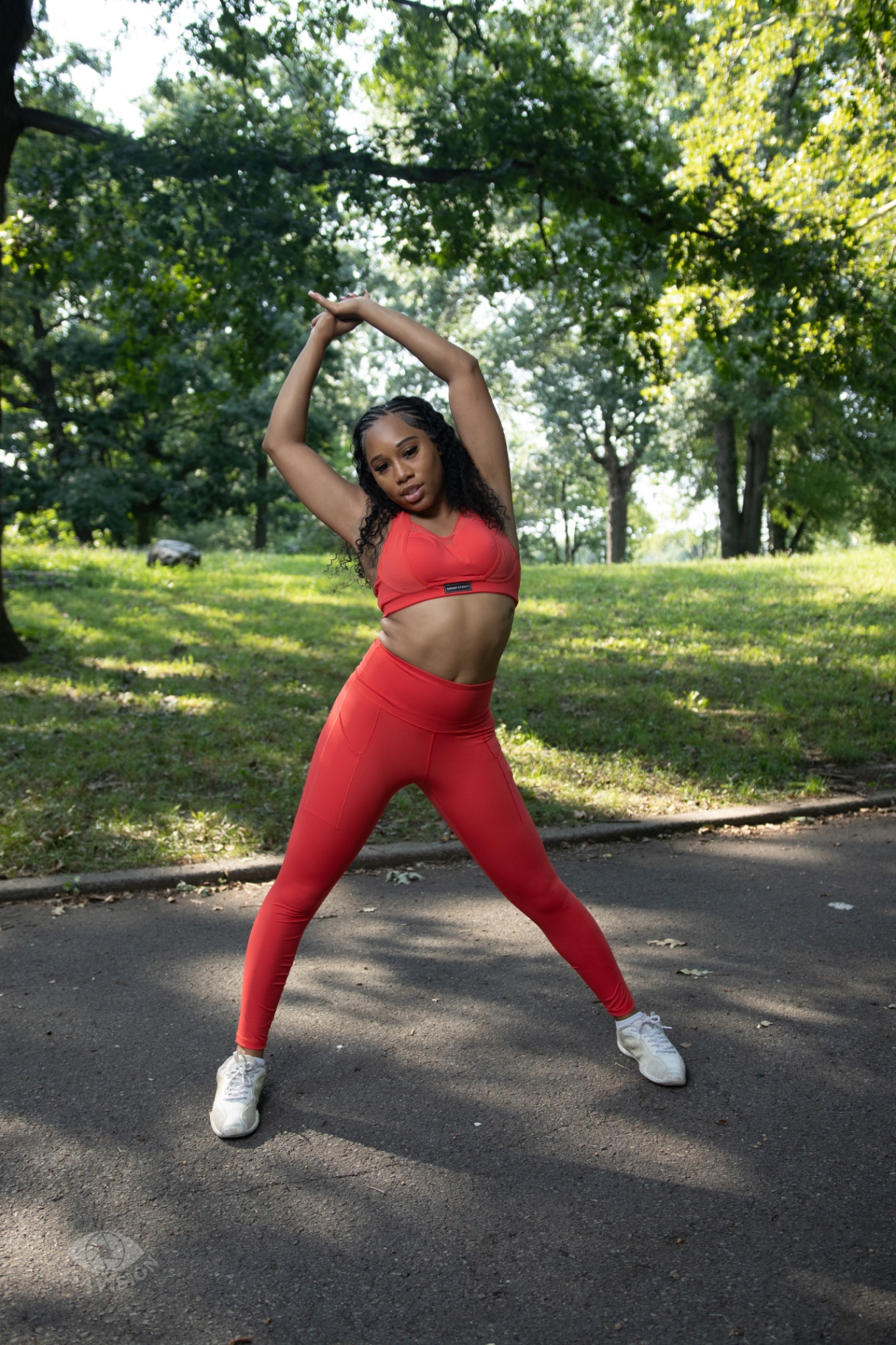 Taniya stretching with arms overhead in a red activewear set in the park