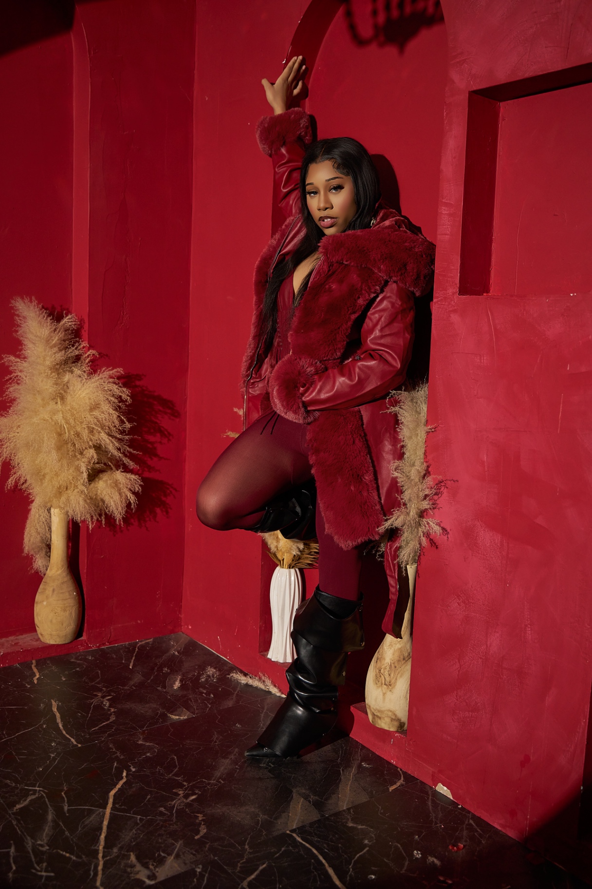Taniya posing in a crimson fur-trim leather coat and black boots inside a dramatic red room